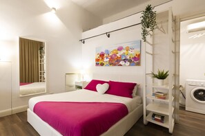 Une chambre moderne avec un grand lit, une étagère murale remplie de livres, un lave-linge et un tableau floral aux couleurs vives.