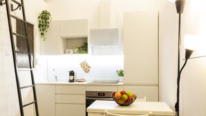 Une cuisine compacte avec des meubles blancs, un lave-vaisselle, un panier de fruits posé sur le comptoir et une échelle appuyée contre le mur.