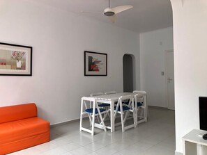 A room with a white ceiling fan, a white table with four chairs, a white sofa, and two framed artworks on the wall.