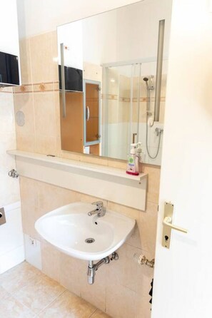 Une salle de bain avec un lavabo blanc, un miroir au-dessus, et un espace douche visible à travers une porte en verre.