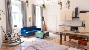 A modern living room with a blue sofa, a rocking chair, a coffee table, and a dining table. There is a kitchen area with cabinets and a stove.