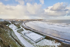 Een kustgebied met een verharde promenade die naar een strand leidt, een omheind sportcomplex en woongebouwen op de achtergrond.