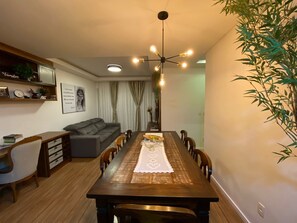 A dining area with a long wooden table, chairs, a chandelier, a sofa, and a plant.