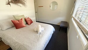 A neatly made bed with pillows, a small stool, a mirror, and a window with blinds.