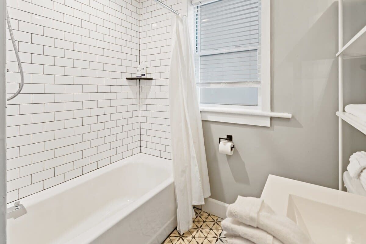 A bathroom with white tiled walls, a bathtub, a window with blinds, and a towel rack.