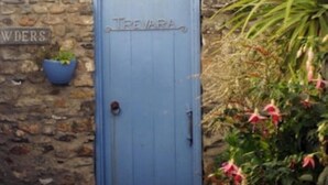 A blue gate with the name "TreVara" on a stone wall, a blue planter with plants, and a view of the sea.