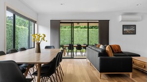 A modern living room with a wooden dining table, black sofas, and a view of a green garden through glass doors.