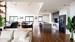 Un salon moderne comprenant un canapé, une table à manger et des chaises, une cuisine équipée d’un réfrigérateur et d’une cuisinière, et de grandes fenêtres laissant entrer la lumière naturelle.