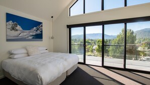 Une chambre dotée d’un grand lit, d’un tableau représentant un paysage de montagne et d’un balcon offrant une vue sur la verdure et les montagnes.