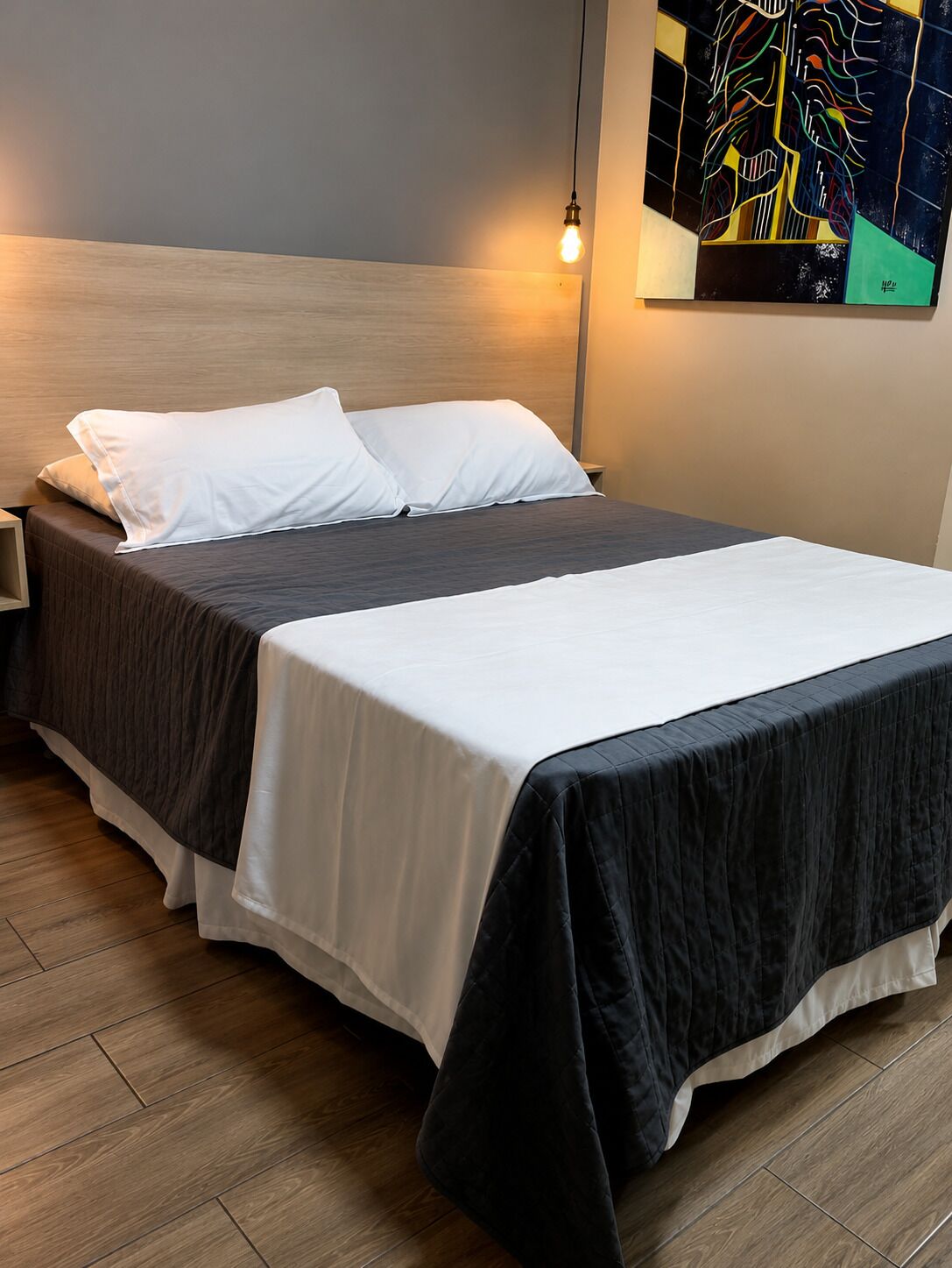 Een net opgemaakt bed met wit en donker beddengoed, een houten hoofdbord en een moderne lamp.