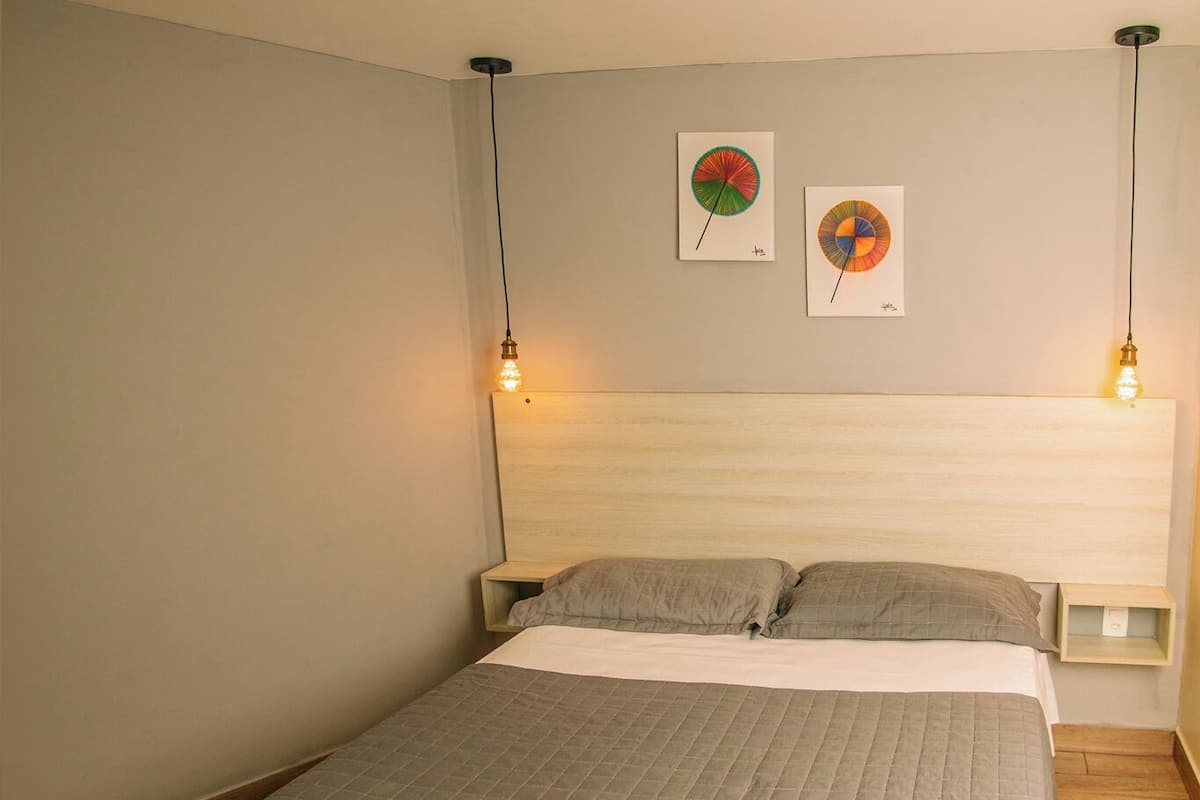 Un lit bien fait, recouvert d’une courtepointe grise matelassée, avec deux appliques murales et deux œuvres d’art encadrées accrochées au mur.