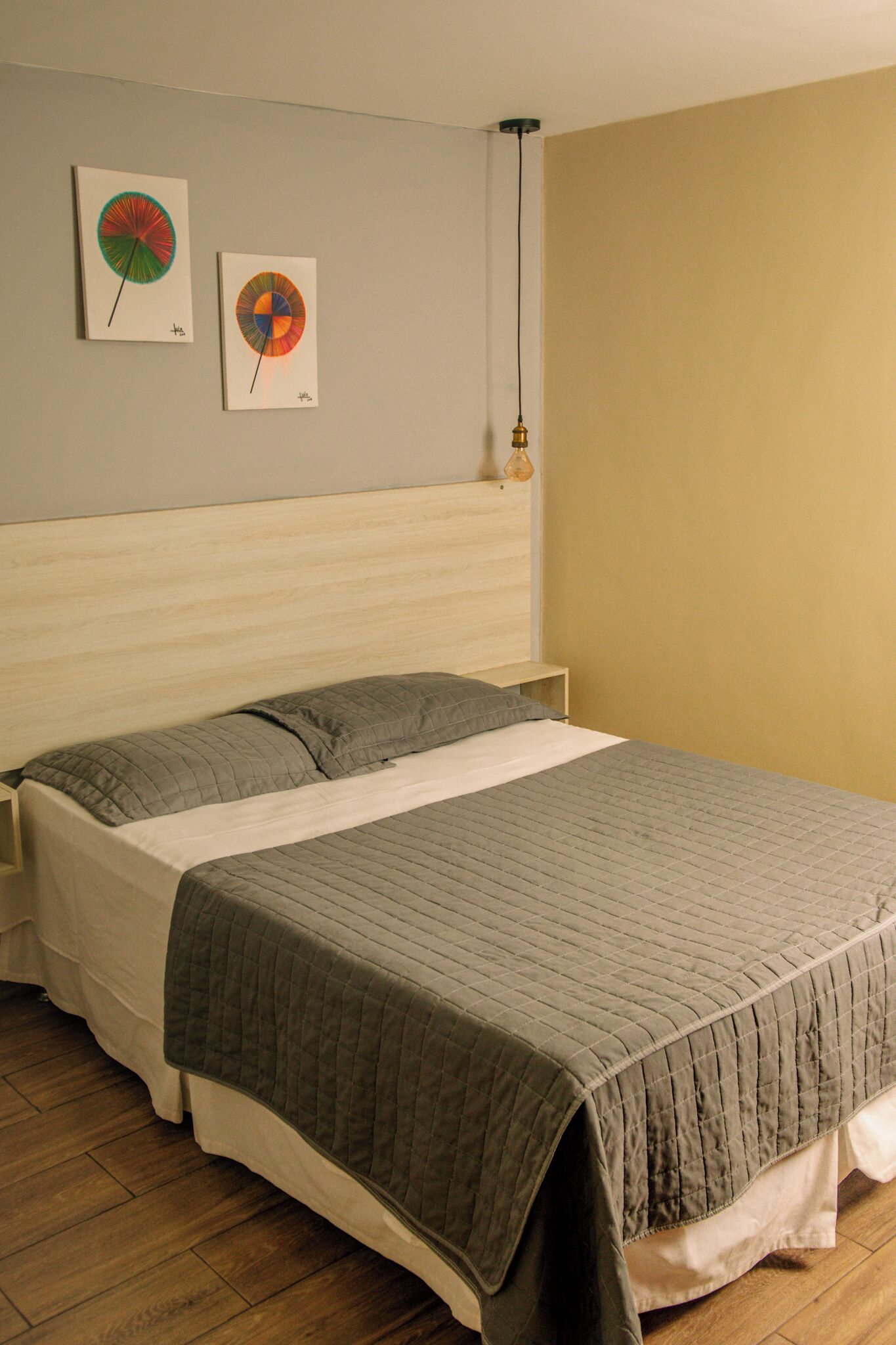 A neatly made bed with a gray quilt, a wooden headboard, and two wall art pieces depicting colorful circular designs.
