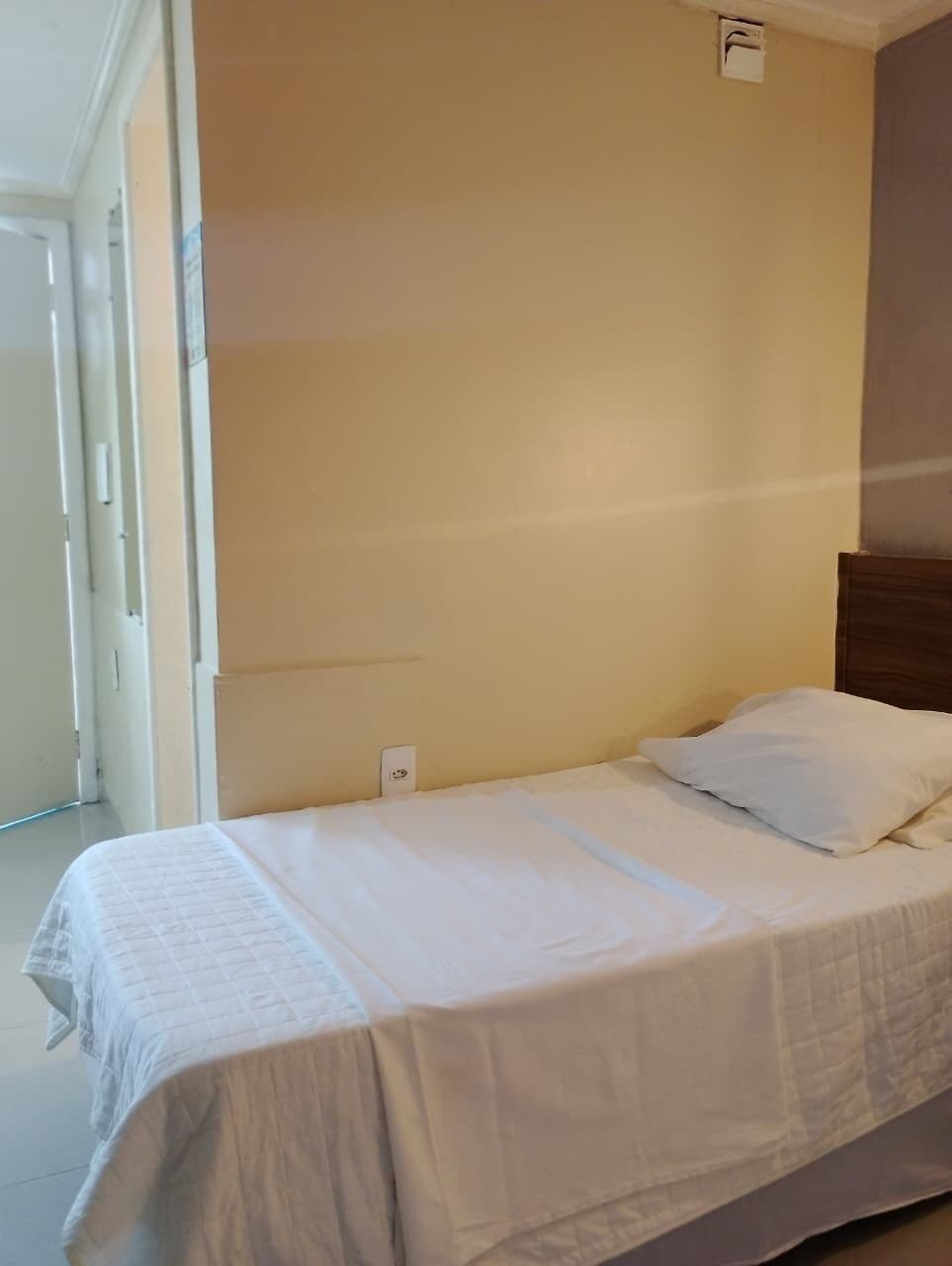 Ein Einzelbett mit weißer Bettdecke und Kissen, ein Holzkopfende und eine schlichte Wand mit Lichtschalter.