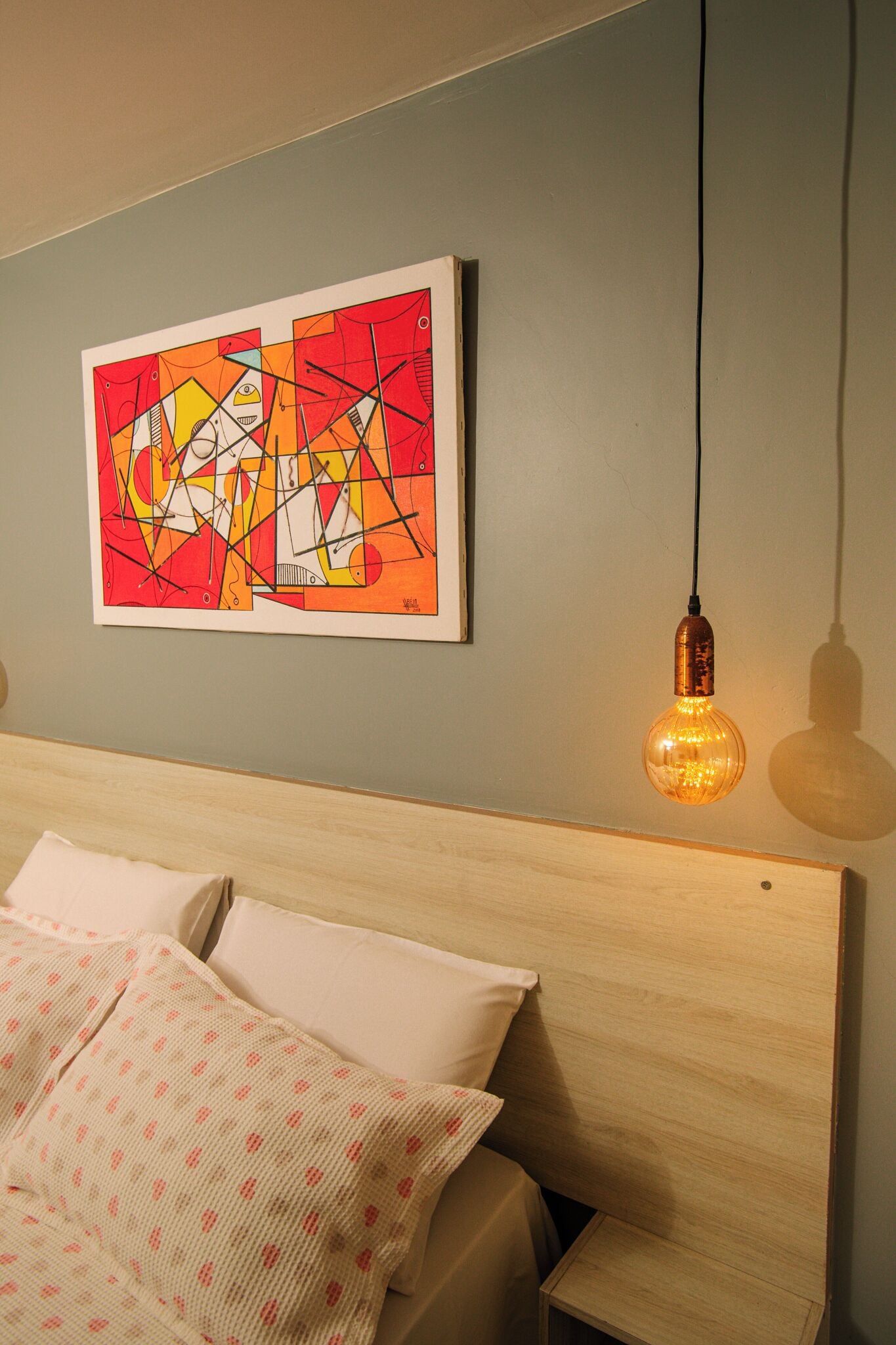 A bedroom with a wooden headboard, a bed with white and patterned bedding, a wall-mounted abstract painting, and a hanging Edison-style light fixture.