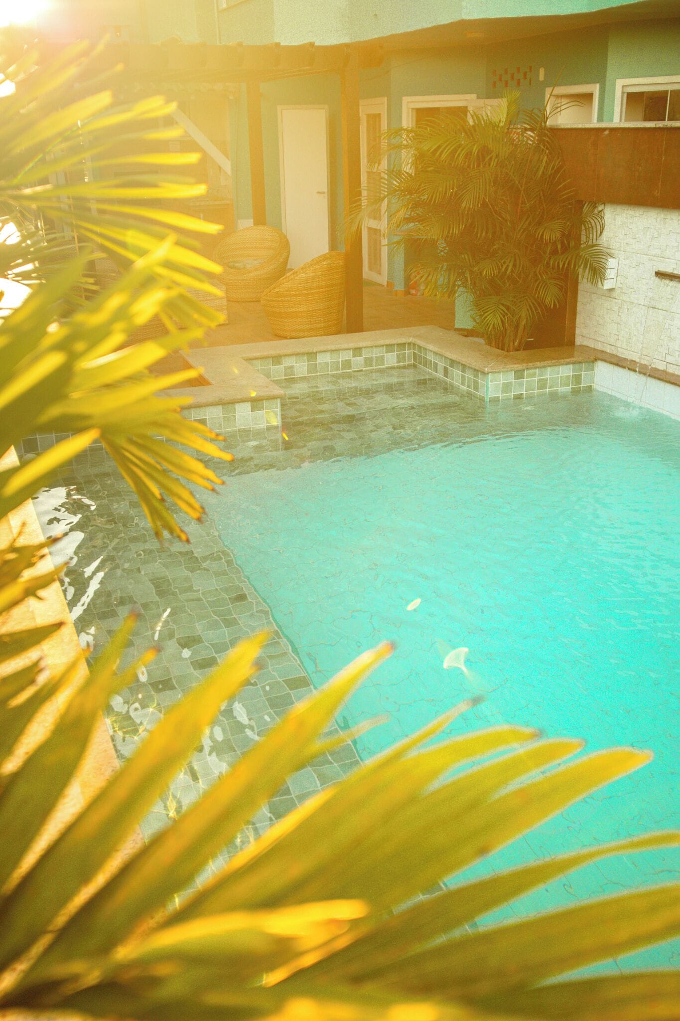 Uma piscina com bordas azulejadas, rodeada por um pátio também azulejado e uma porta que dá para uma varanda coberta.