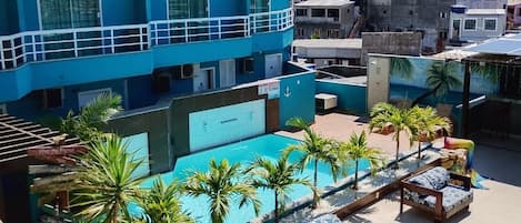A rooftop pool area with lounge chairs and a yellow umbrella, surrounded by a blue building with multiple floors.