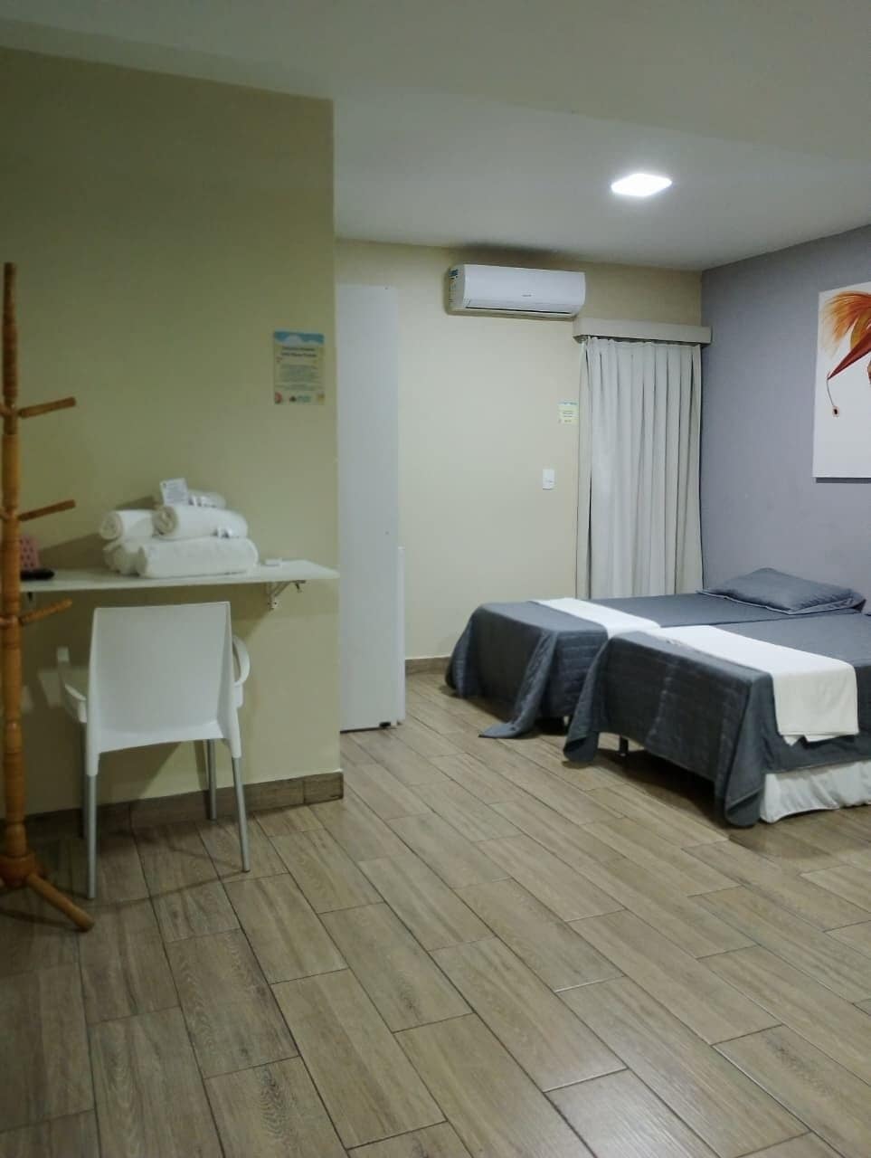 Quarto com duas camas, uma escrivaninha com cadeira, aparelho de ar condicionado e janela com cortinas.