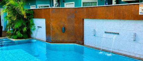 A swimming pool with a wooden deck, a water fountain, and a multi-level building with air conditioning units.