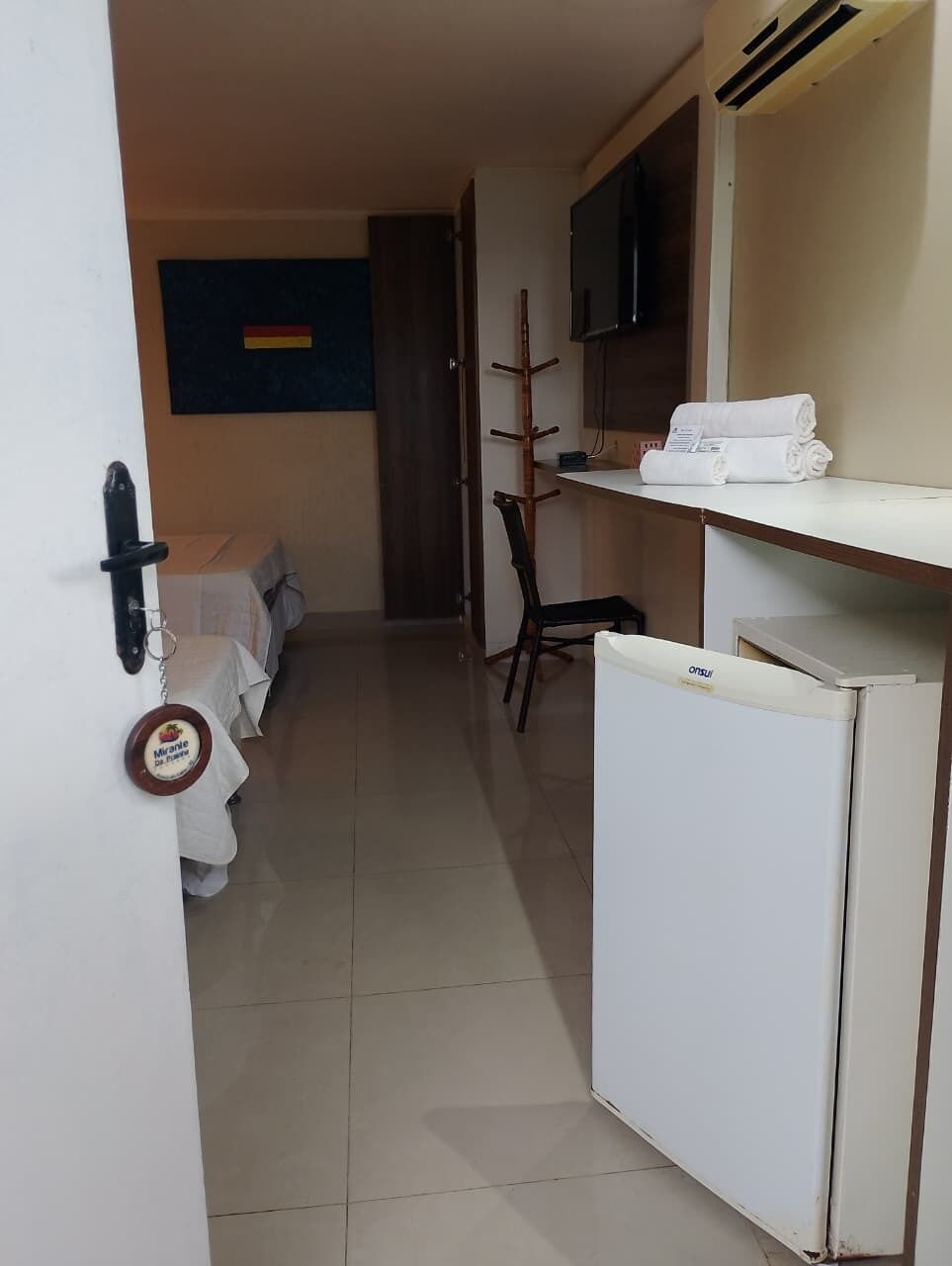 Habitación de hotel con una cama, una silla, un refrigerador pequeño, un televisor montado en la pared y un perchero.