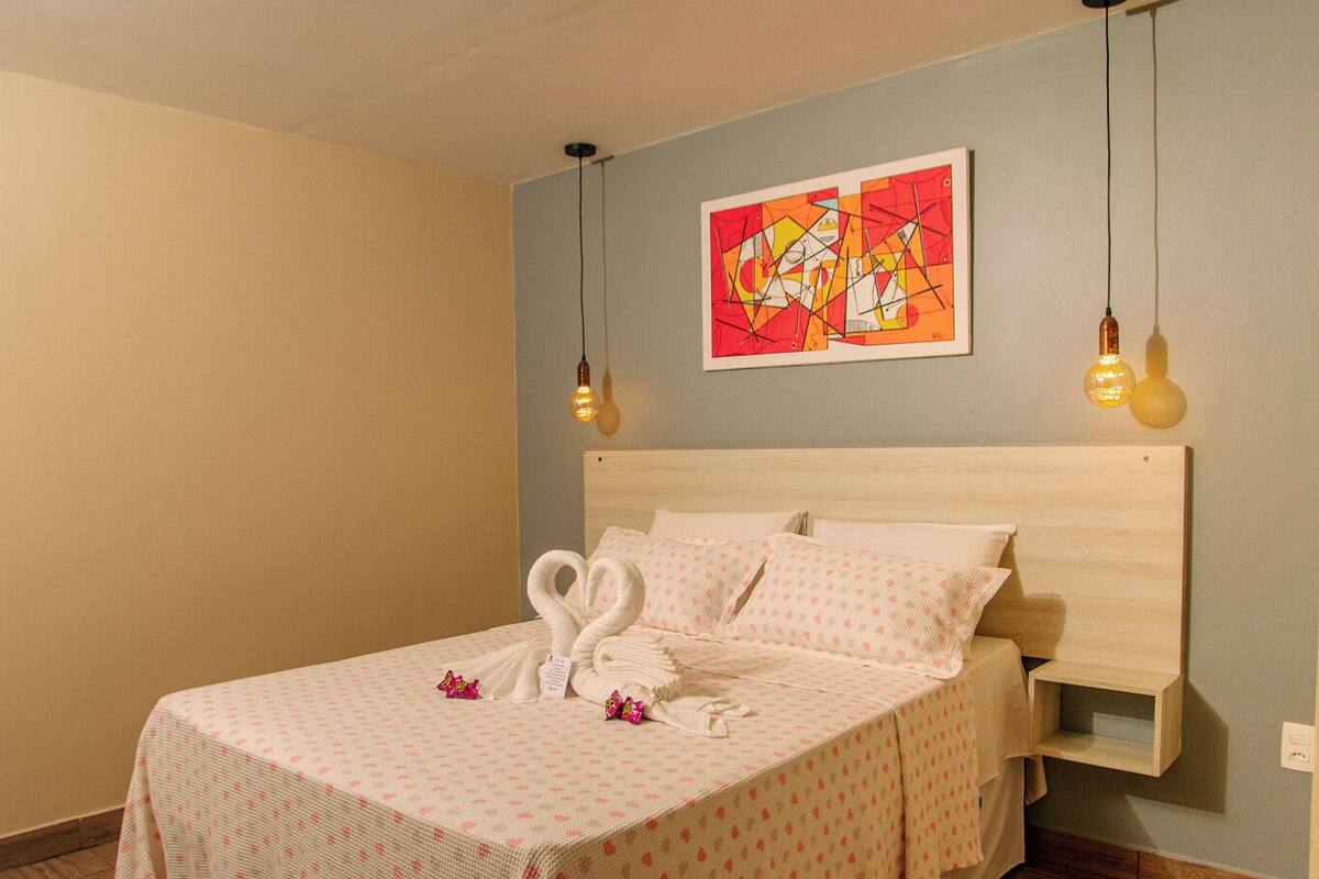 Un lit bien fait, recouvert d’une couvre-lit blanche et rose à motifs, avec une tête de lit en bois et deux appliques murales.