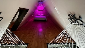 Een smalle gang met een bed aan het einde, verlicht met paars licht, en een hangmat aan de rechterkant.