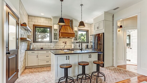 A modern kitchen with white cabinets, a large island with stools, hanging pendant lights, and a brick-patterned floor.