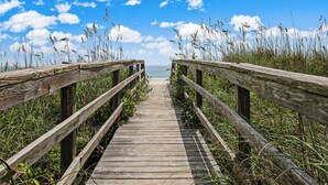 Ein hölzerner Steg führt zu einem Strand mit blauem Himmel und Wolken.