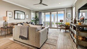 Ein geräumiges Wohnzimmer mit einem großen Sofa, einem Couchtisch und einem Essbereich mit Holztisch und Stühlen. Es gibt einen Deckenventilator, ein großes Fenster mit Blick und eine Pflanze in der Ecke.