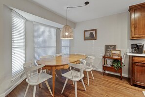 A dining area with a wooden table and white chairs, a hanging light fixture, and a small shelf with decorative items.