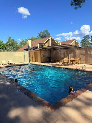 A swimming pool with a covered patio area, a fenced yard, and a building with a sloped roof.