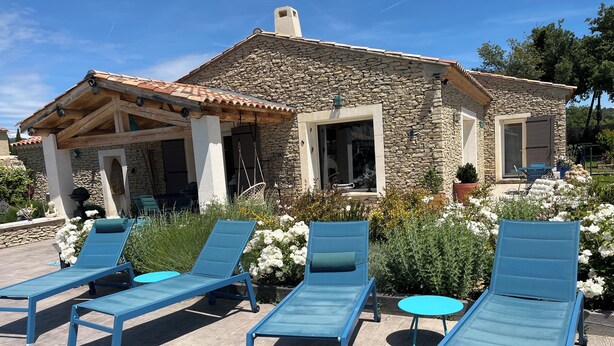 A stone house with a tiled roof, a covered patio with blue lounge chairs, and a garden with white flowers. - Villa Rube's (Gordes)