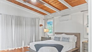 A bedroom with a wooden bed frame, white bedding, a blue rug, and a window with curtains. - Surfers Paradise: Ocean View, Walking Distance to Sunset Beach NUC:1990/NUC-2073 (Haleiwa)