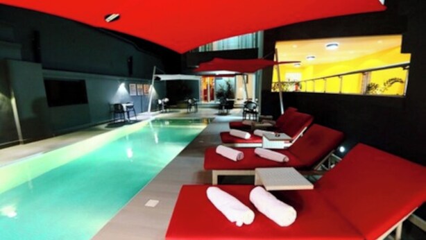 Zona de piscina con sillas reclinables rojas y sombrillas, suelo de baldosas y pared de fondo amarilla.