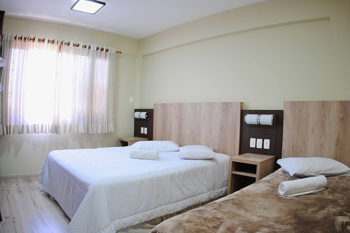 Quarto de hotel com duas camas, cabeceira de madeira, mesa de cabeceira com abajur e janela com cortinas.