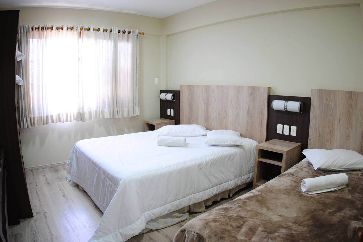 Quarto de hotel com uma cama grande, duas mesas de cabeceira, uma cabeceira de madeira e uma janela com cortinas.