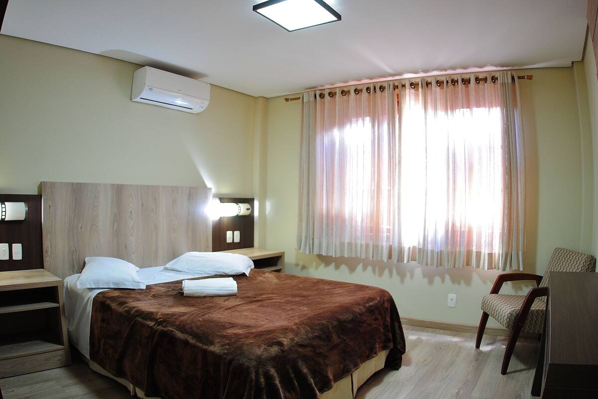 Quarto de hotel com cama, mesas de cabeceira, uma cadeira e janela com cortinas.