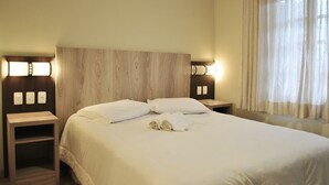 Habitación de hotel con cabecera de madera, dos mesitas de noche con lámparas, cama con ropa blanca y ventana con cortinas translúcidas.