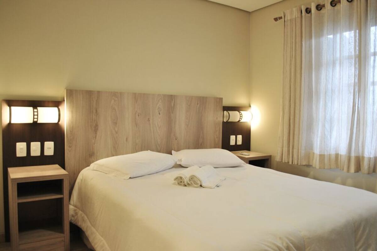 Quarto de hotel com cabeceira de madeira, duas mesas de cabeceira com luminárias, cama com roupa de cama branca e uma janela com cortinas leves.