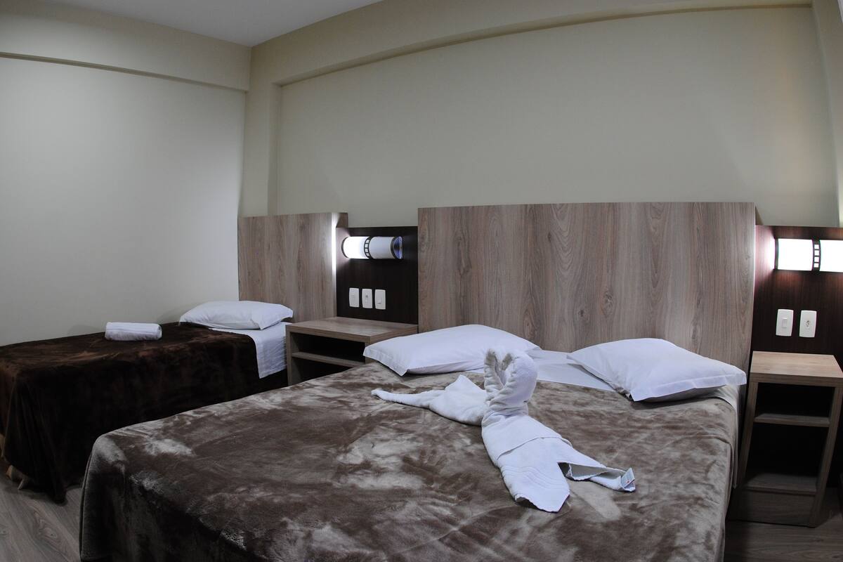 Quarto de hotel com duas camas de solteiro, uma cama de casal e uma cabeceira de madeira.