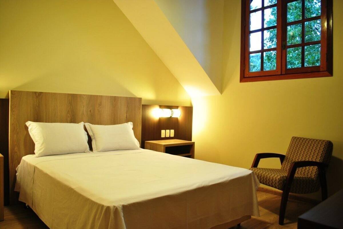 Quarto com cama, mesa de cabeceira, luminária, cadeira e janela com vista para árvores.