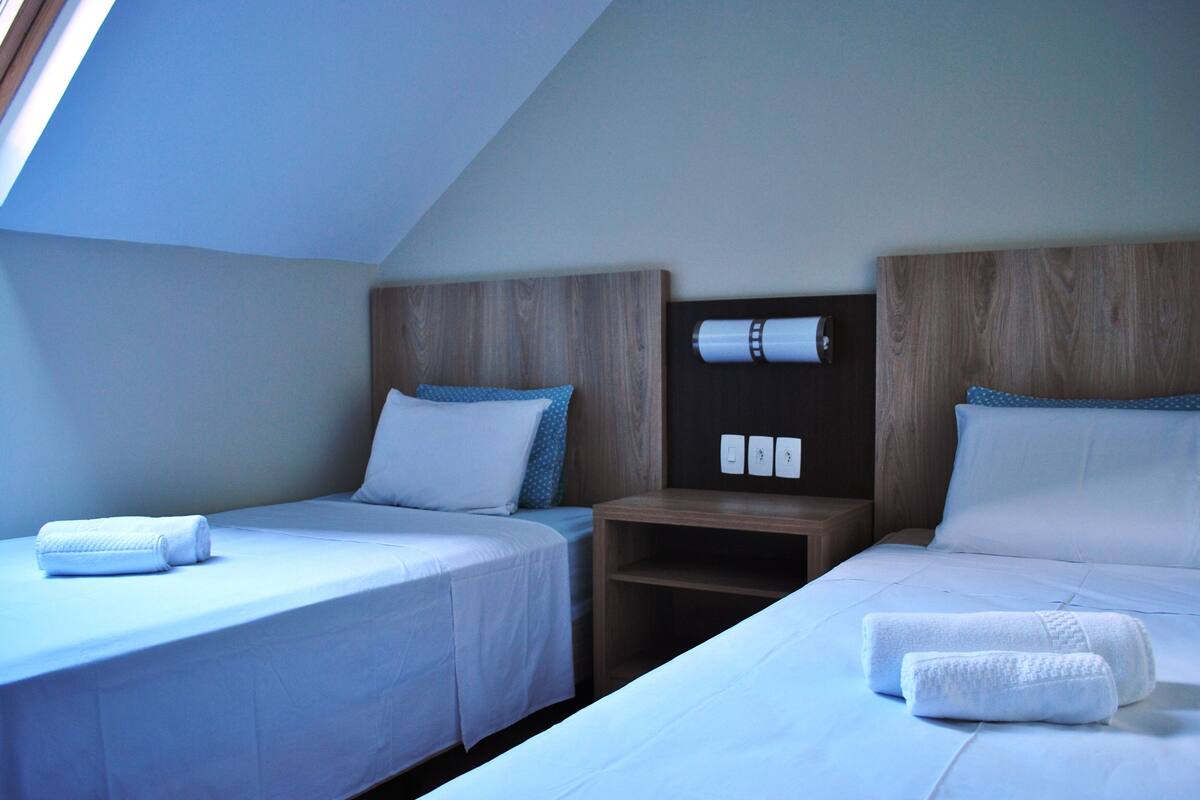 Quarto de hotel com duas camas de solteiro, cabeceira de madeira, mesa de cabeceira e claraboia.