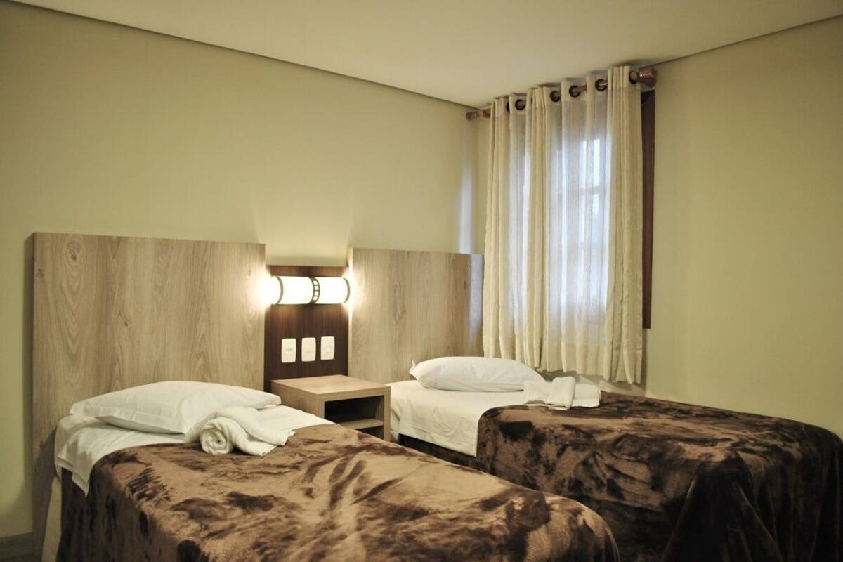 Quarto de hotel com duas camas, cabeceira de madeira, mesa de cabeceira e janela com cortinas.