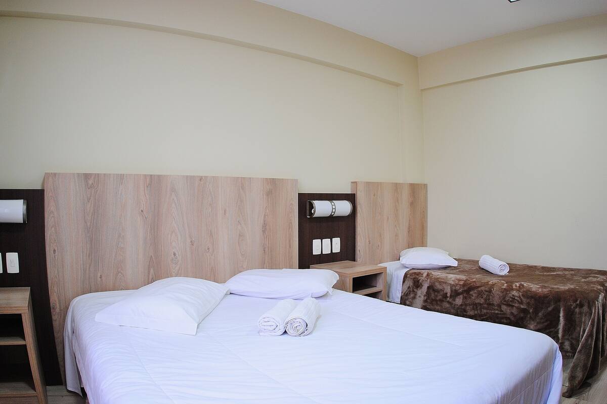 Quarto de hotel com duas camas, uma cabeceira de madeira, uma mesa de cabeceira pequena de madeira e uma TV fixada na parede.