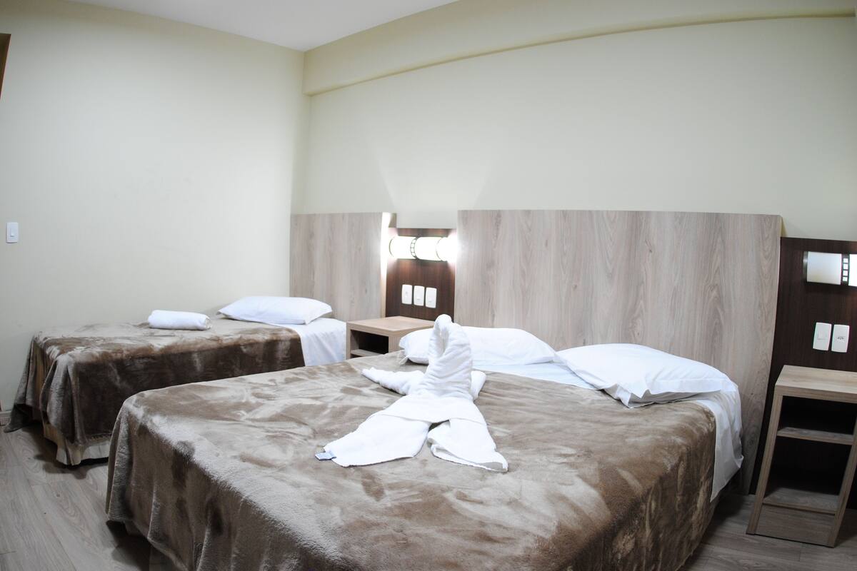 Quarto de hotel com duas camas, cabeceira de madeira, mesa de cabeceira e luminária fixada na parede.