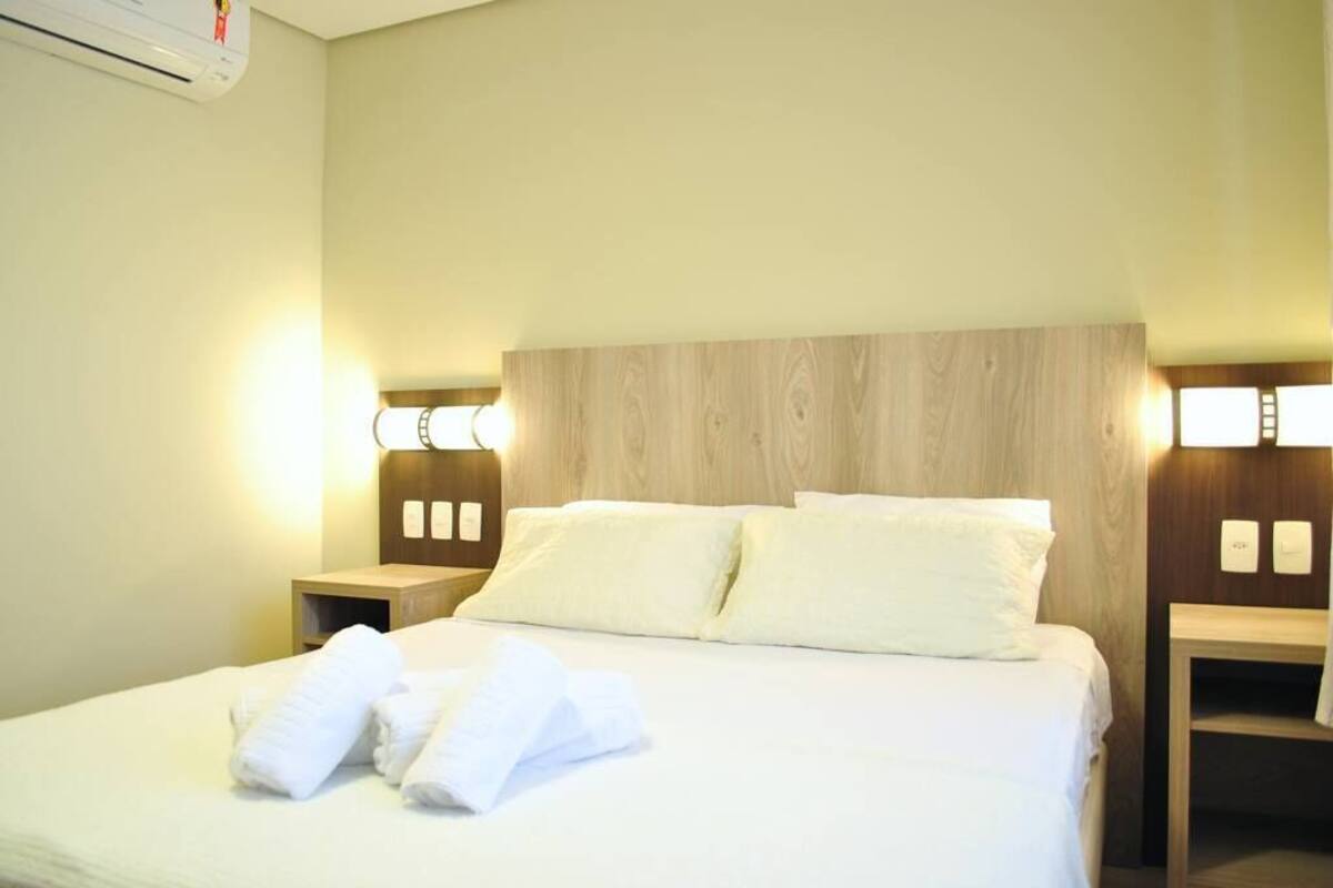 Cama bem arrumada com lençóis e toalhas brancas, cabeceira de madeira e abajures laterais.