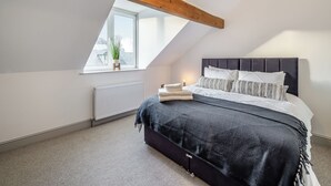 Ein Schlafzimmer mit schrägen Wänden, einem Bett mit dunklem Kopfteil, einem Nachttisch mit Lampe und einem Fenster mit Pflanze.
