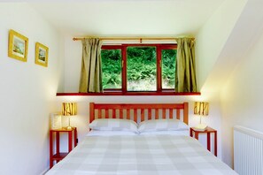 Un dormitorio con una cama, dos mesitas de noche con lámparas, una ventana con cortinas y dos cuadros en la pared.