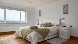 Ett sovrum med ett stort fönster, en säng med kuddar, ett sängbord med en lampa och ett element.