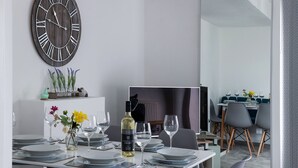 A modern dining room with a large wall clock, a dining table set for four, and a view into another room. - Rivage - Two Bedroom Cottage, Sleeps 4 (Deganwy)