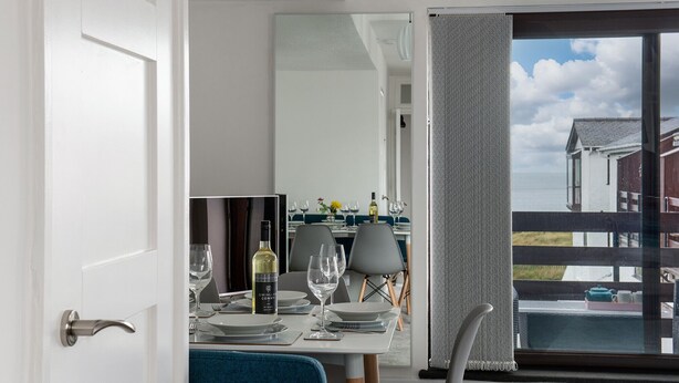 A dining area with a table set for two, a bottle of wine, and a view of the sea through a large window. - Rivage - Two Bedroom Cottage, Sleeps 4 (Deganwy)